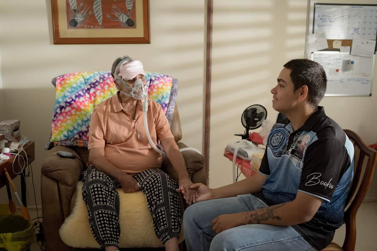 Image of a woman sitting on a couch with a breathing device over her mouth holding hands with a younger man who is caring for her