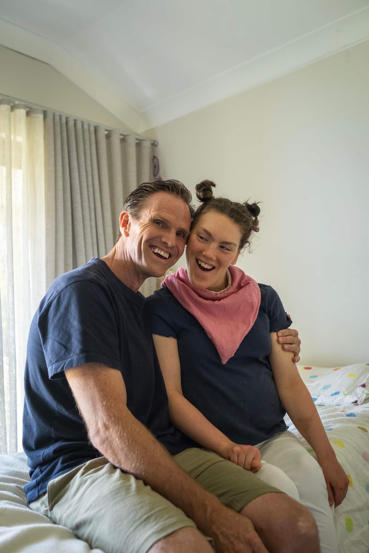 Image of a man wearing a blue tshirt and khaki shorts with his arm wrapped around his daughter who has her hair in two buns on top of her head, a pink scarf around her neck and a navy tshirt. They are both smiling widely.