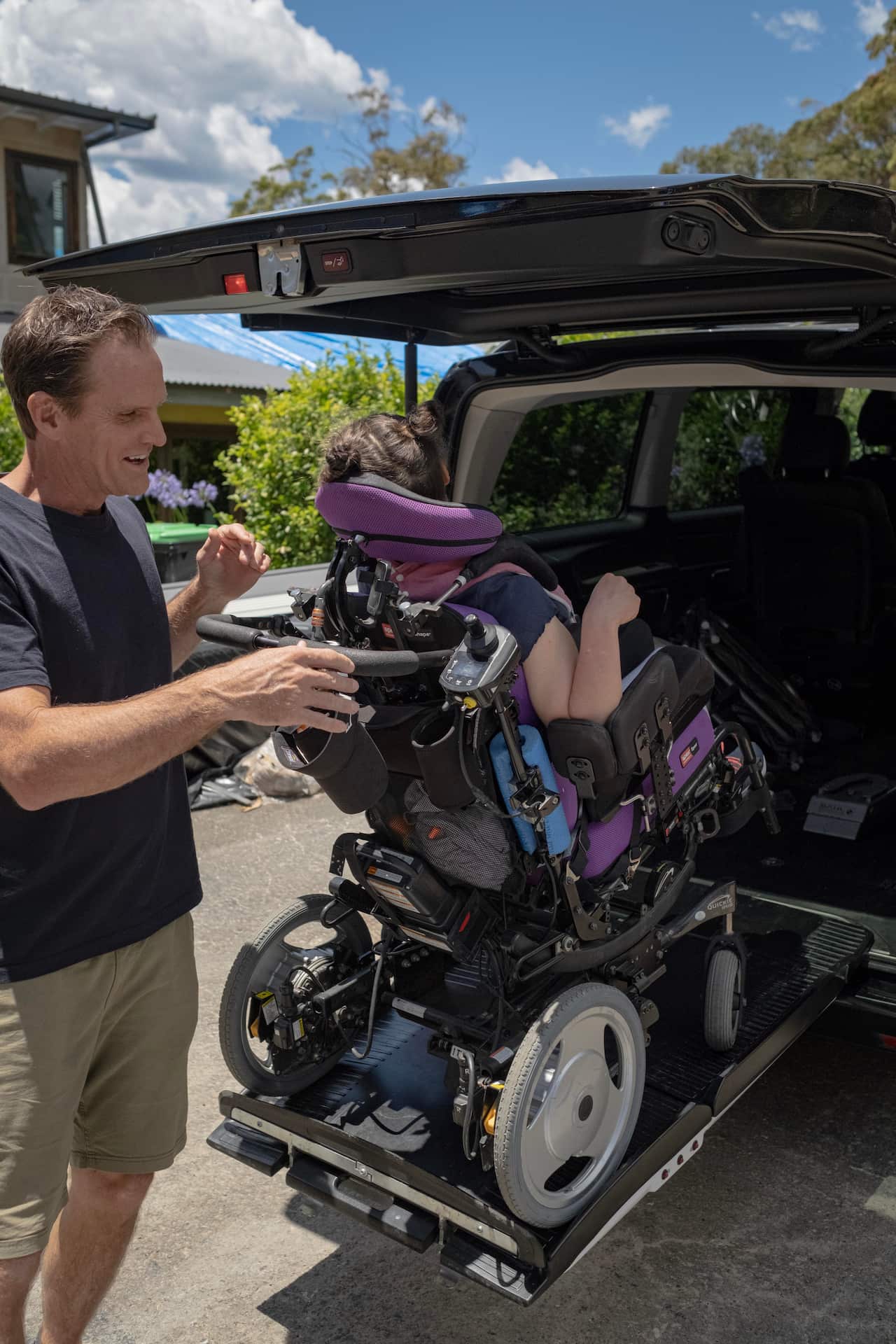 Image of a man using a lift at the back of the car to move his daughter in a wheelchair into the back of a large car.