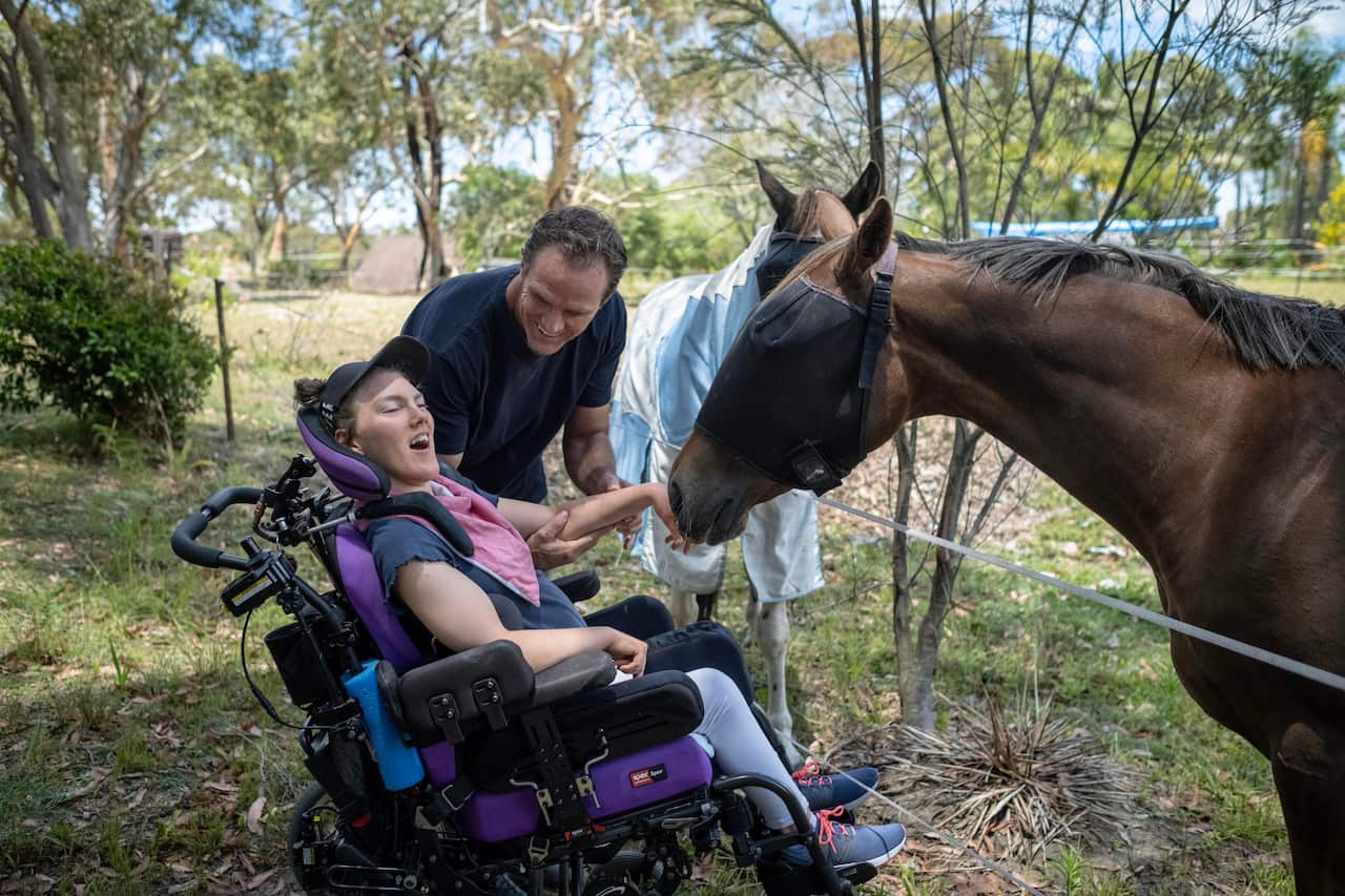 Image of a man in a navy tshirt next to his daughter in a wheelchair. He is holding her hand up to a horse out in a green field.