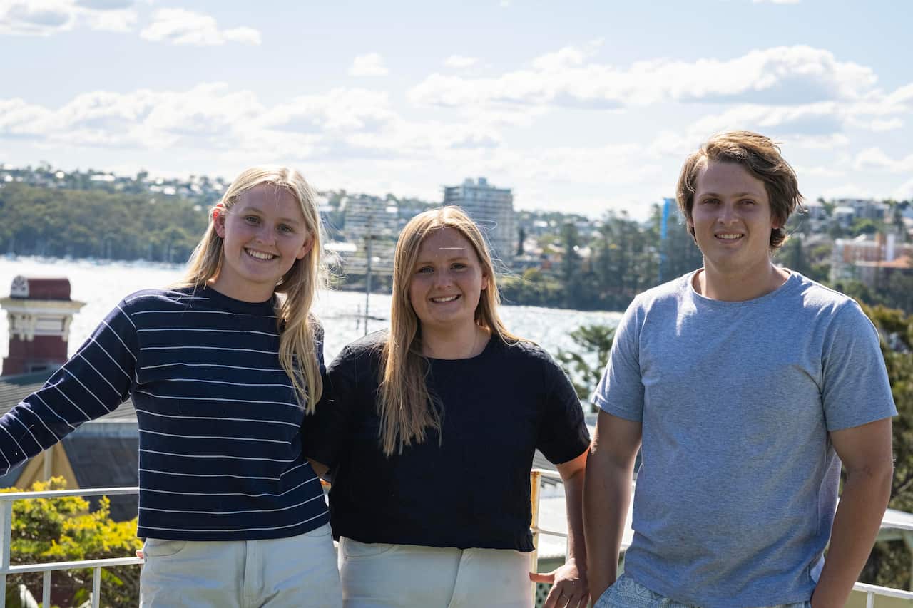 Image of two young women with blonde hair standing next to a young man with brown hair on a balcony in the sun.