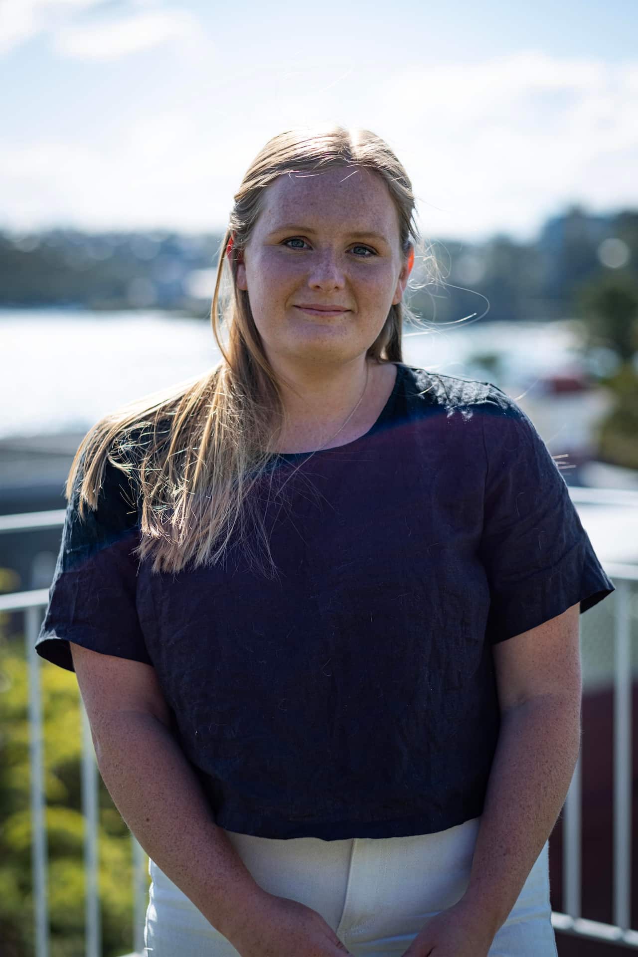 Image of a young woman wearing a navy top and white pants standing outside on a balcony in the sun.