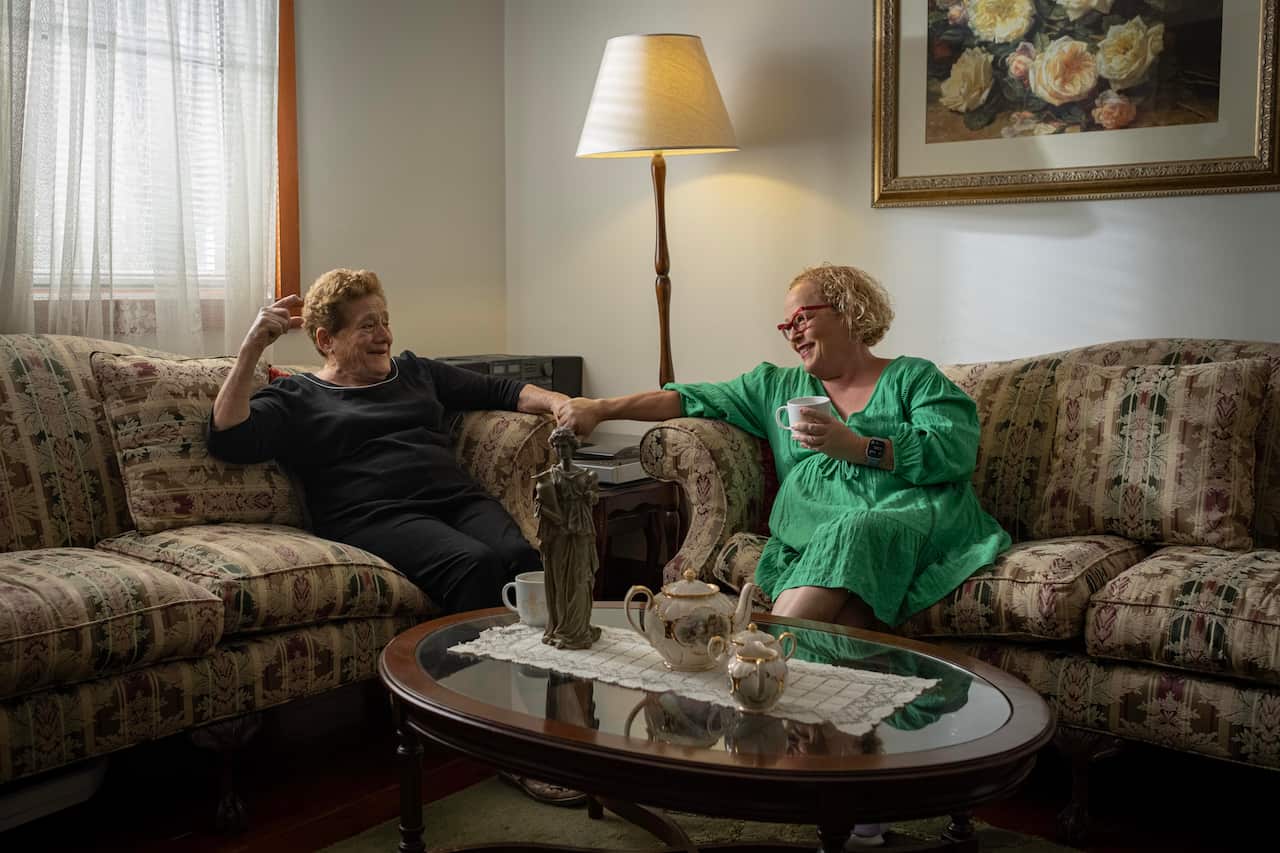 Image of a woman wearing black and a woman in a green dress holding hands and speaking animatedly in a cozy loungeroom.