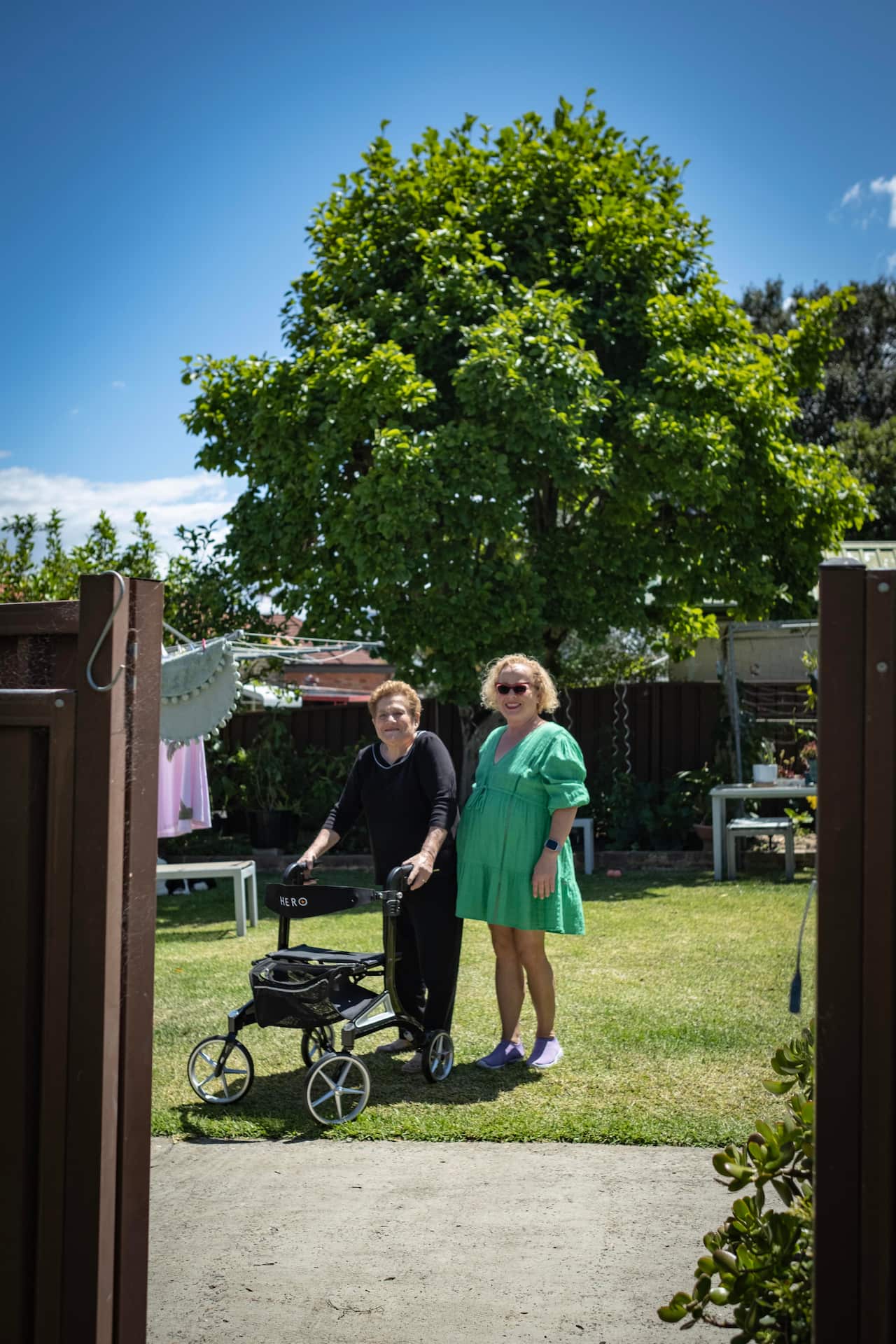 Image of a woman in black with a walker standing next to a woman in a green dress both standing in a sun drenched garden near a hills hoist and large tree. 