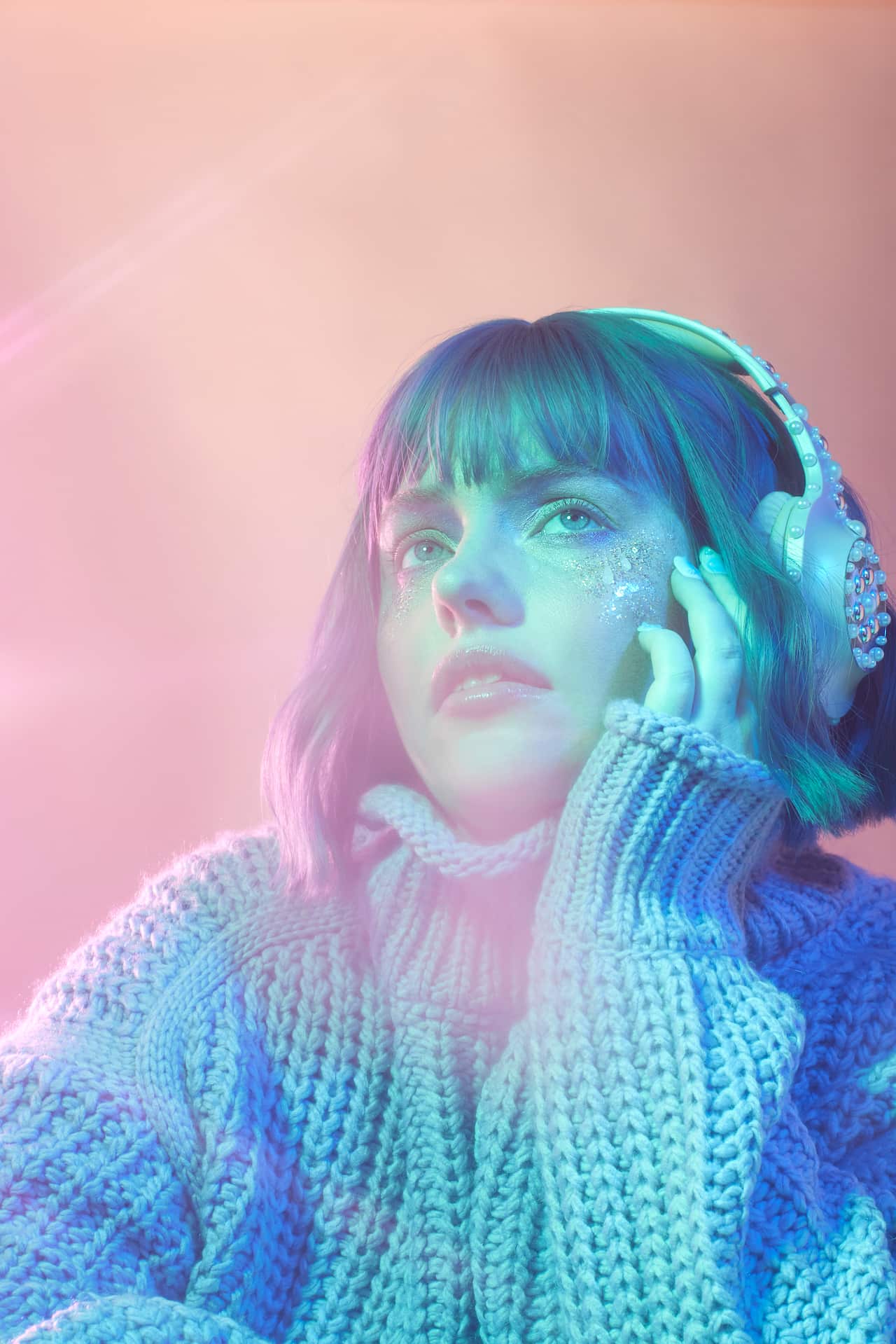 A woman with purple hair and pink bedazzled headphones looking into the distance in front of a pink background. 