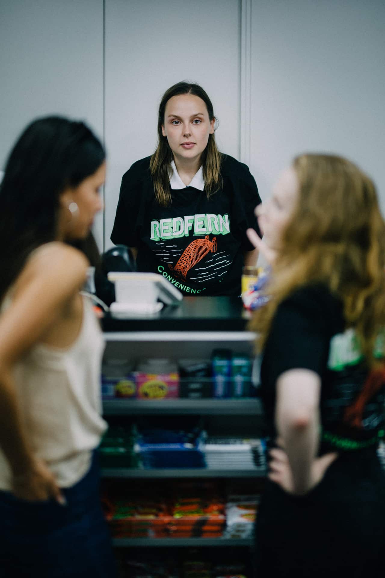 Woman facing camera wearing a black printed tee over a white collared shirt. She is behind a counter, watching two other women in the foreground.