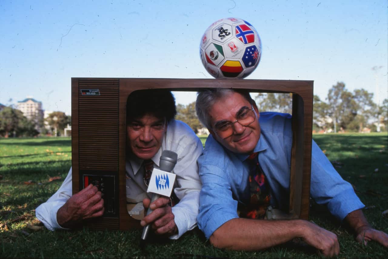 Two men posing for a photo. They are laying on the ground at an oval and are sticking their heads through a prop that looks like a television. One is holding a microphone and there is a soccer ball on top of the prop.