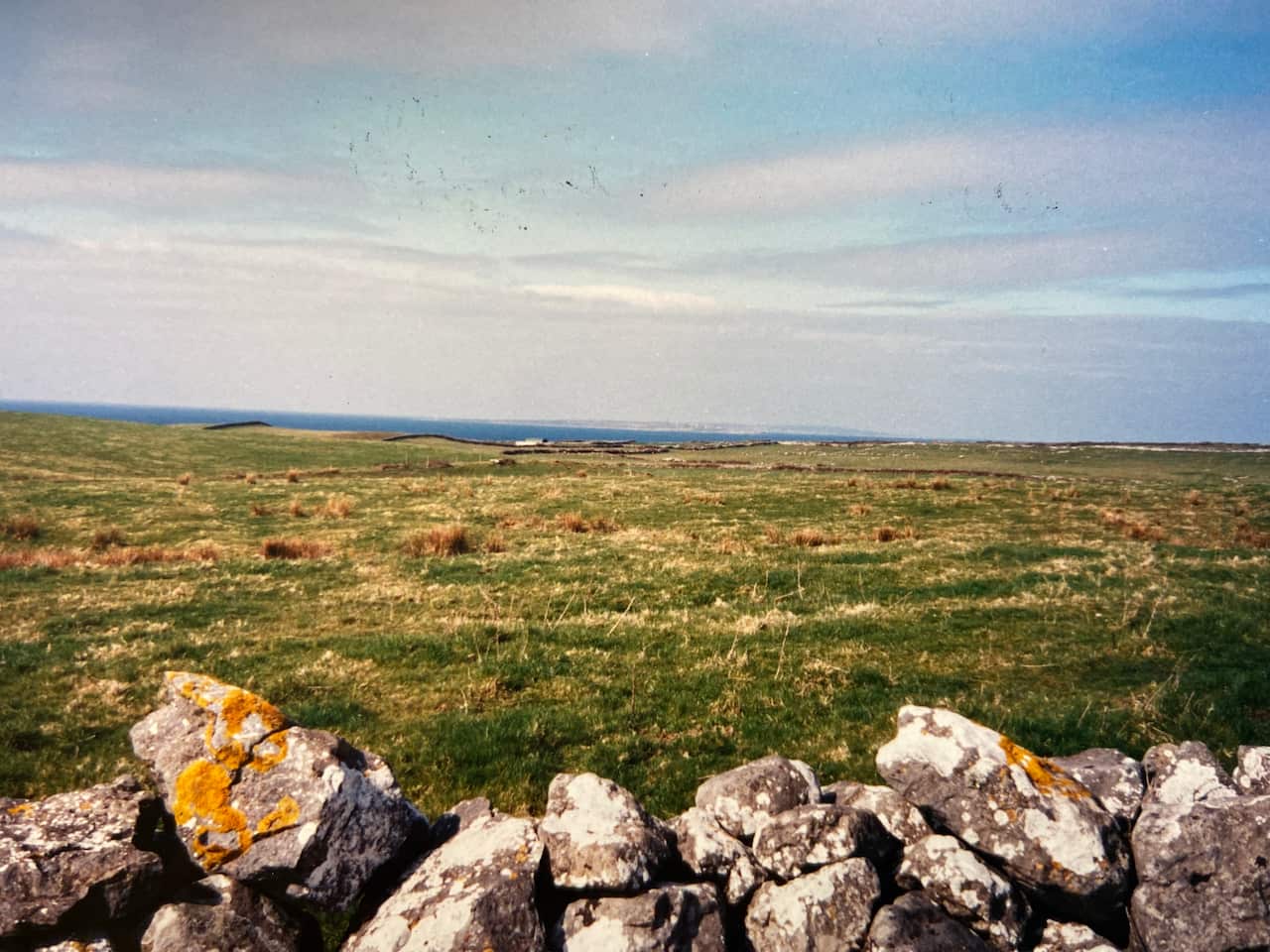 A landscape; there is a row of grey rocks in the foreground, green grass in the midground and a sliver of ocean visible on the horizon. The sky above is streaked with clouds. The photograph appears aged. 