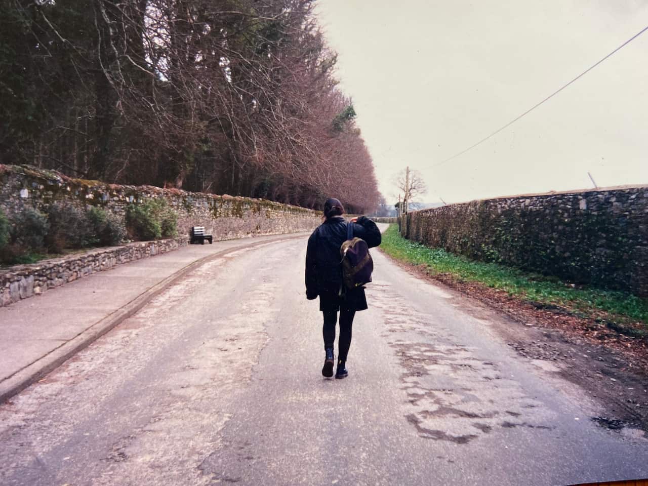 A woman with a backpack slung over her shoulder, walking down a road on a cloudy day. On the right side of the road, there is a hedge and greenery. On the left side of the road is a row of trees with bare branches behind a stone wall.