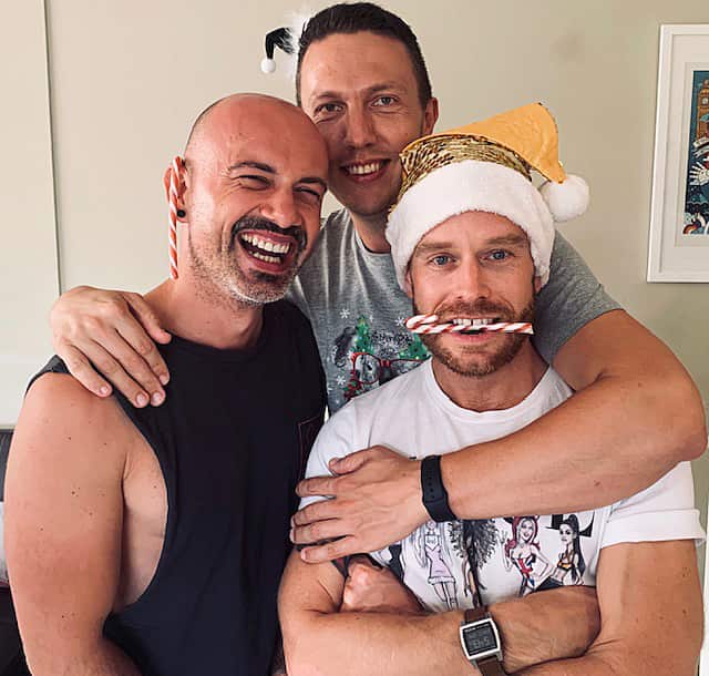 Three men embracing and smiling at camera, one with a santa hat and candy cane