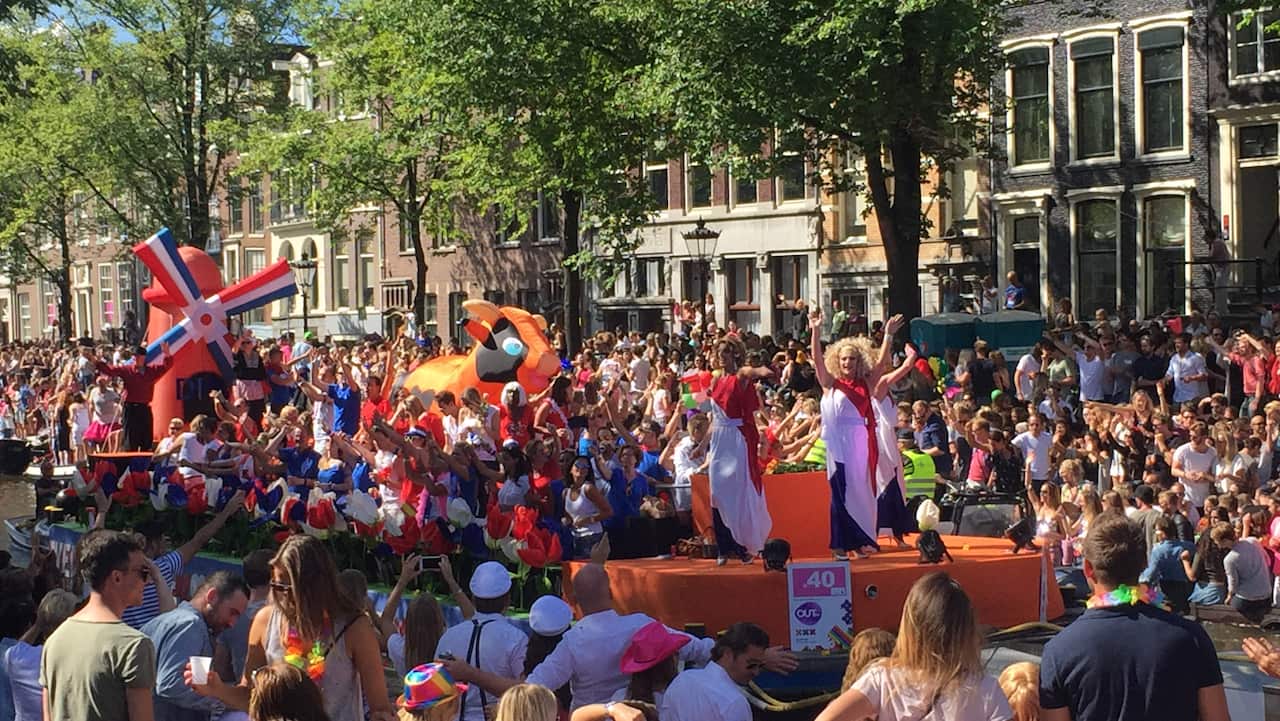 An Amsterdam pride boat motors down the canal, a drag queen is at the head of the boat and an inflatable dutch windmill in orange is at the back. 