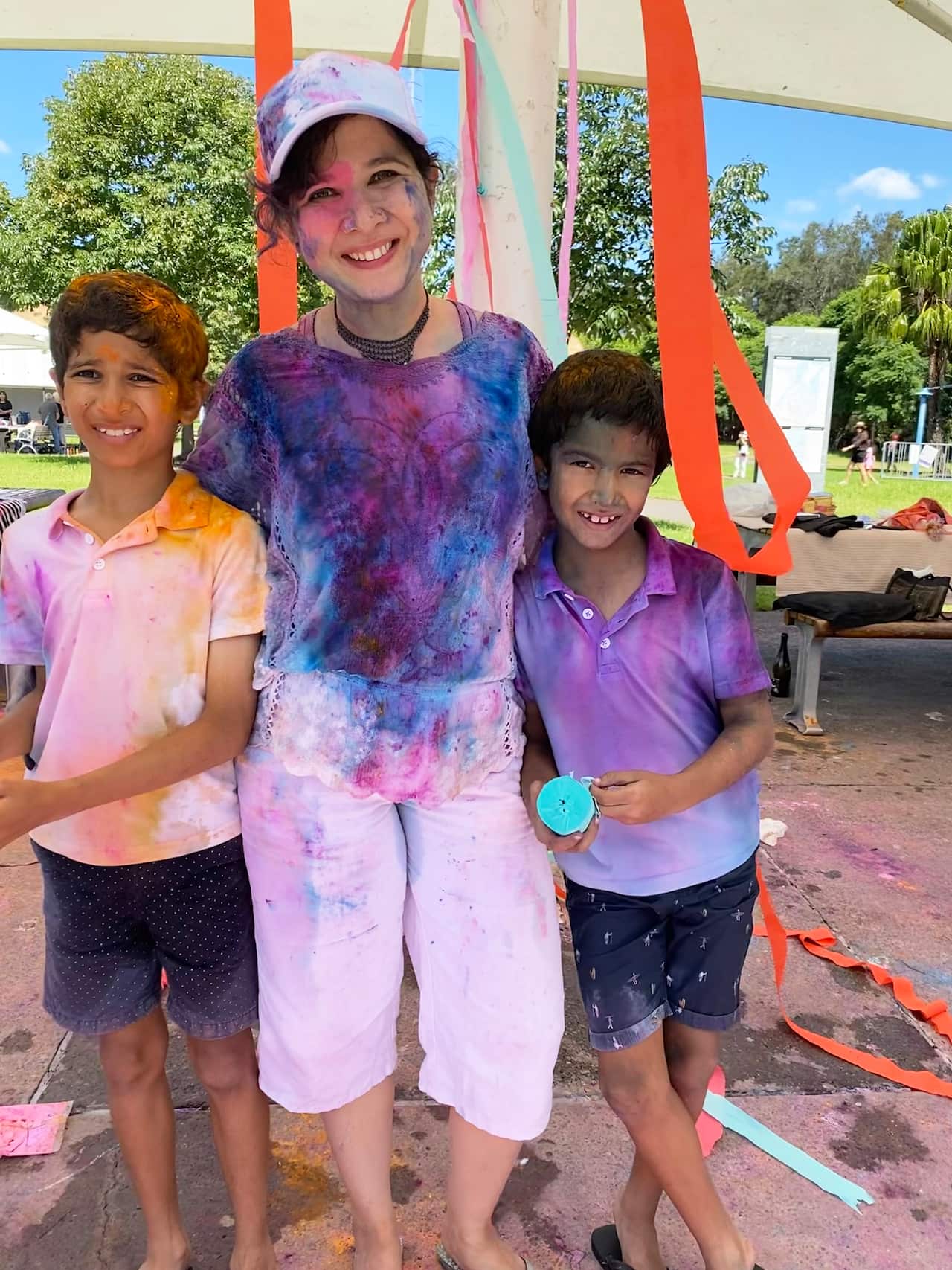 South Asian mother in playground smiling with two boys, covered in colourful powder