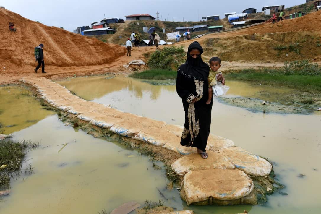 A Rohingya woman carries a child over sandbags through water lying on the ground at the Balukhali refugee camp in Bangladesh on December 11, 2017.