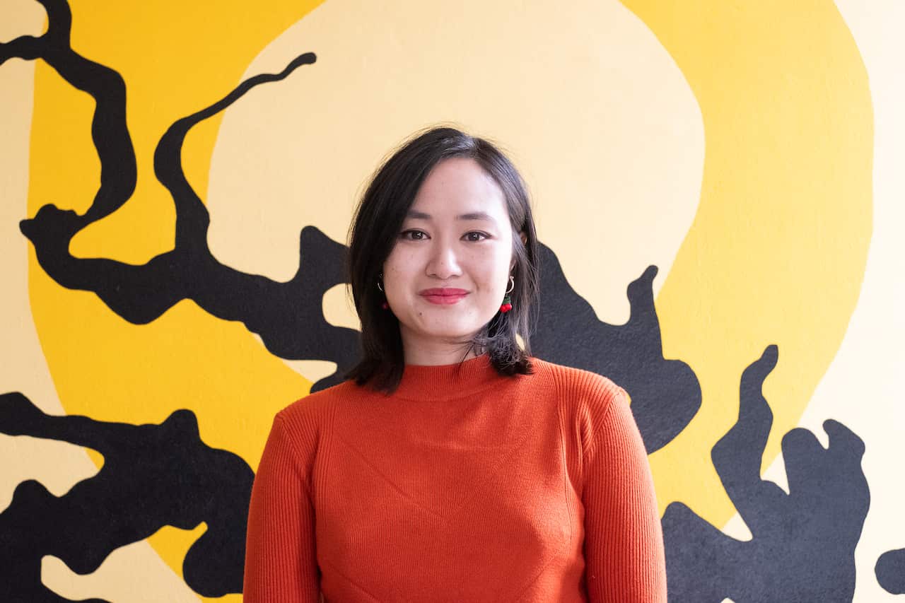 Young Asian woman in red top in front of black and yellow background, slightly smiling at camera