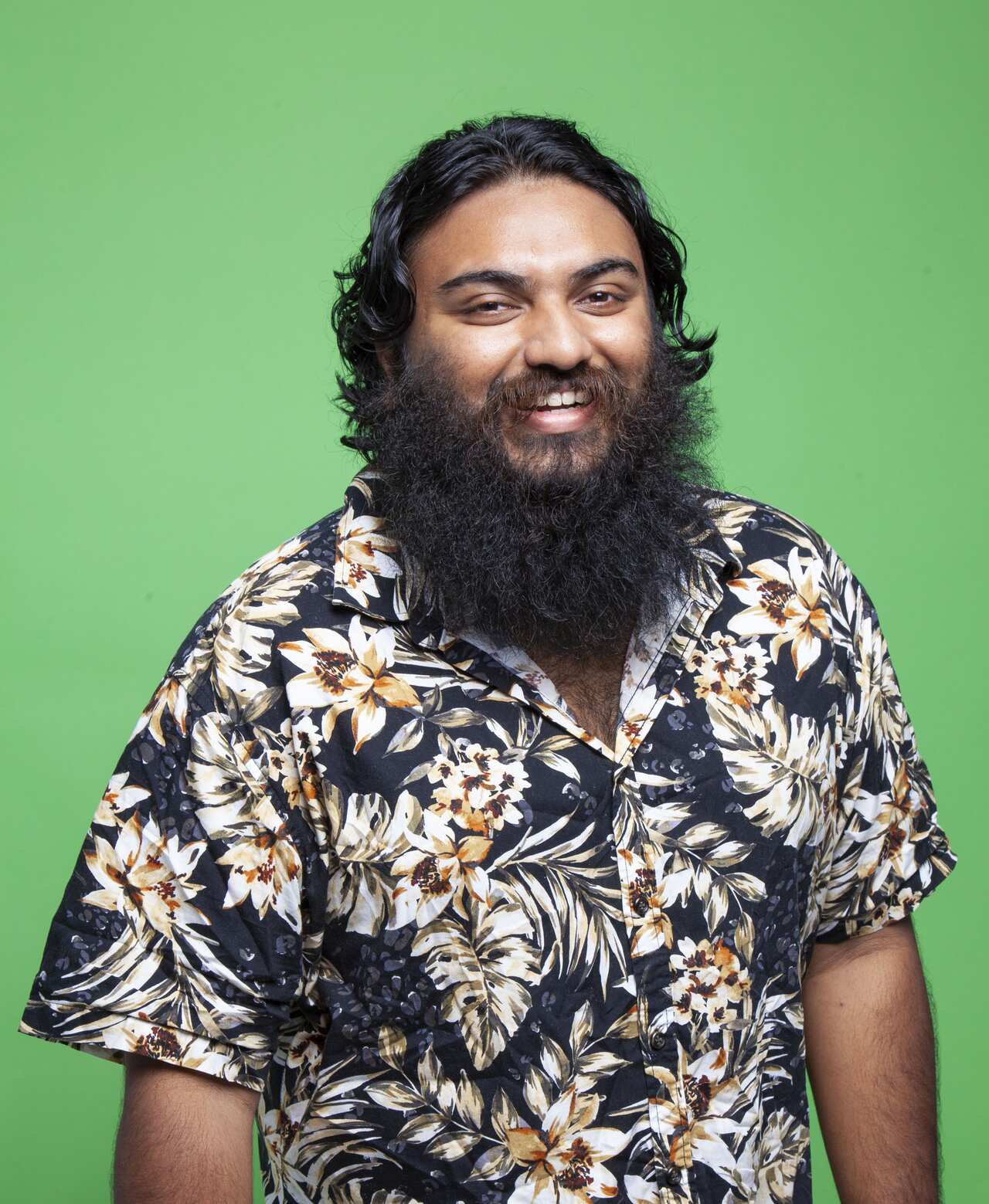 South Asian man with bushy beard wearing a Hawaiian-print shirt, standing in front of a green background and smiling. 