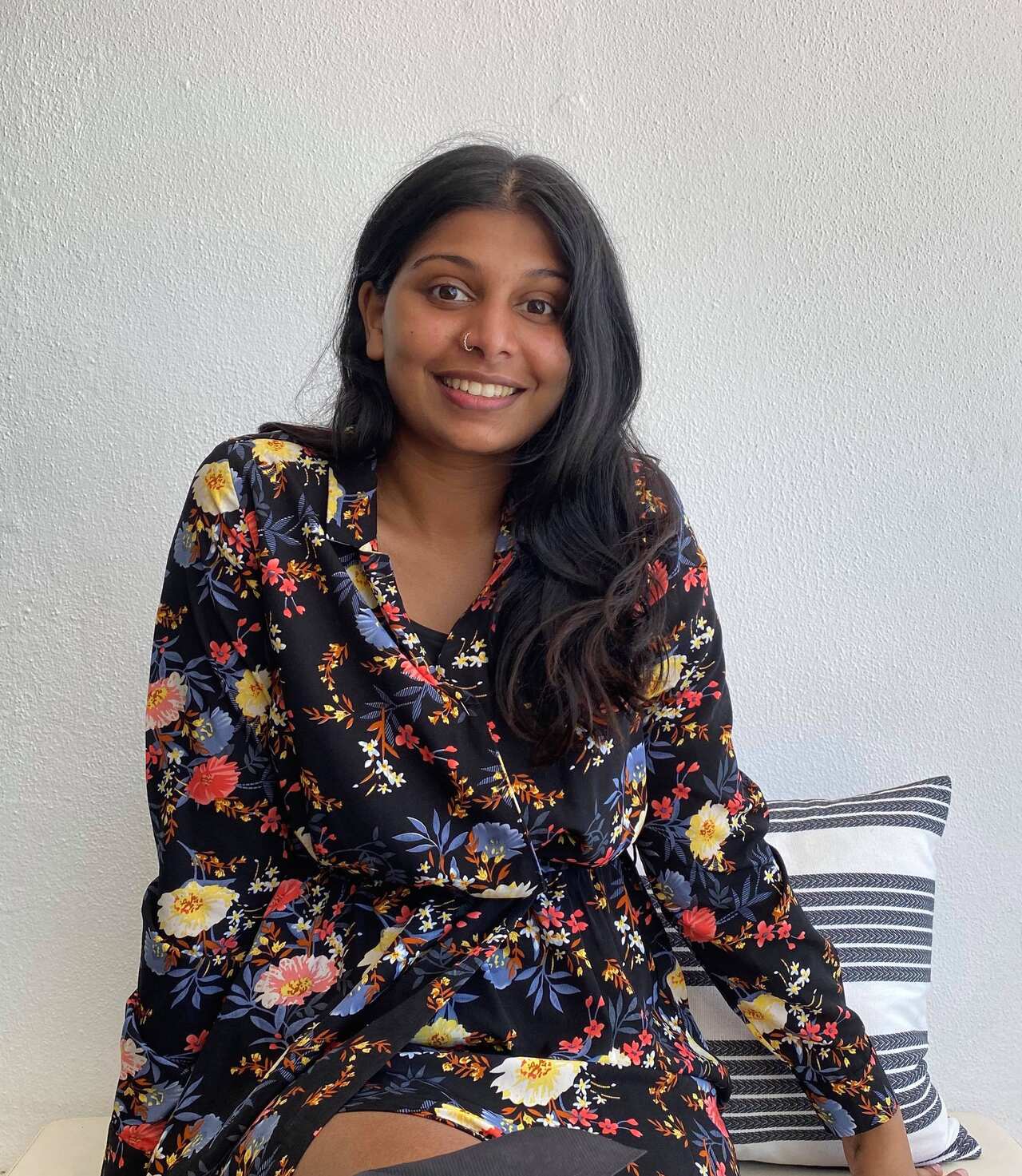 Varuna Naicker wearing a black, red, purple and yellow floral printed dress. She is smiling at the camera and sitting in front of a blank grey wall and striped cushion. 