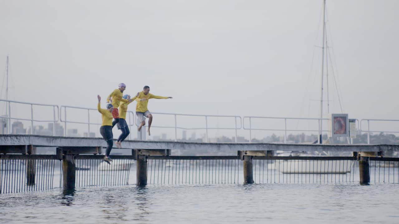 Four lifeguards in yellow rash shirts jumping from a jetty into the ocean. 