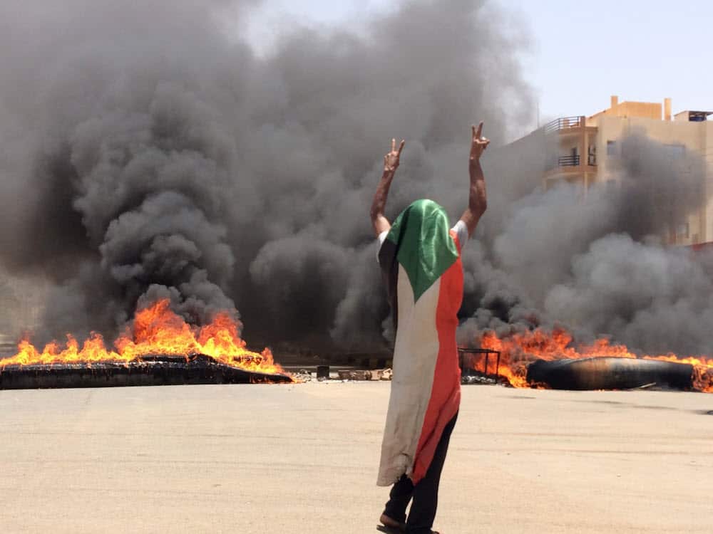 A protester wearing a Sudanese flag in front of burning tires and debris on road 60, near Khartoum's army headquarters