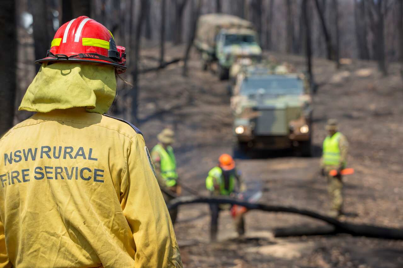 A NSW Rural Fire Service member monitors conditions as Australian Army personnel clear fallen trees near Cobargo.