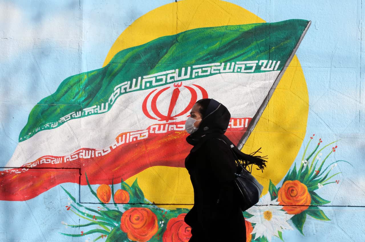 An Iranian woman wearing a mask walks next to a wall painting of Iranian national flag in Tehran, Iran.