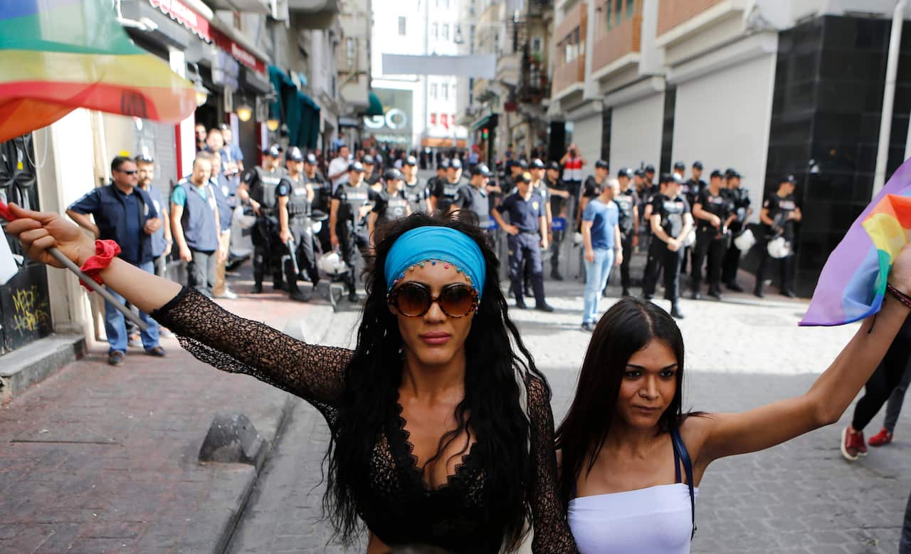 two women holding pride flags stand in front of dozens of riot police on a narrow street. 
