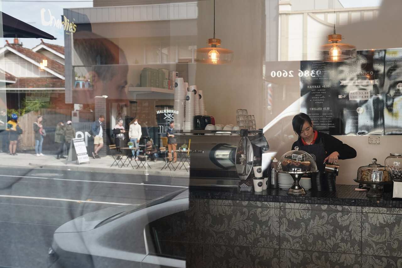 A barista makes coffee in a Brunswick cafe while across the road people queue outside a Centrelink office.