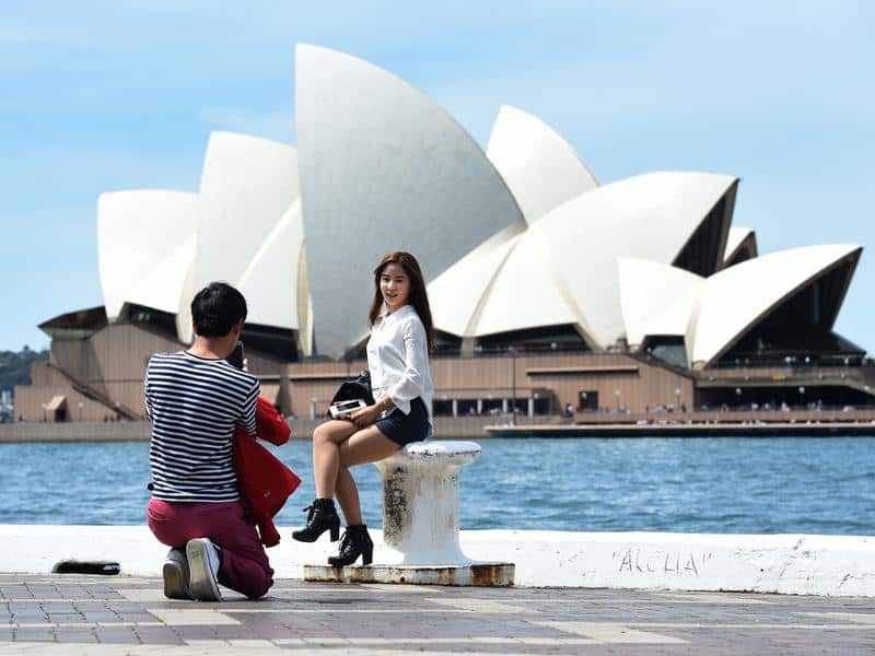 A man takes a photograph of a woman front of the Sydney Opera House