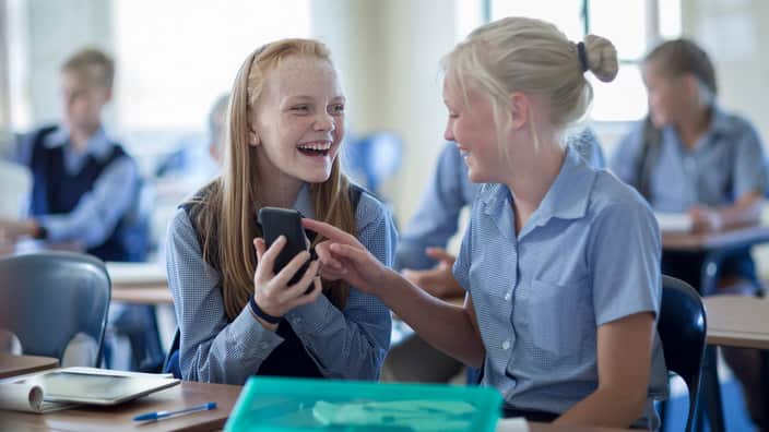 Two happy schoolgirls in classroom with cell phone