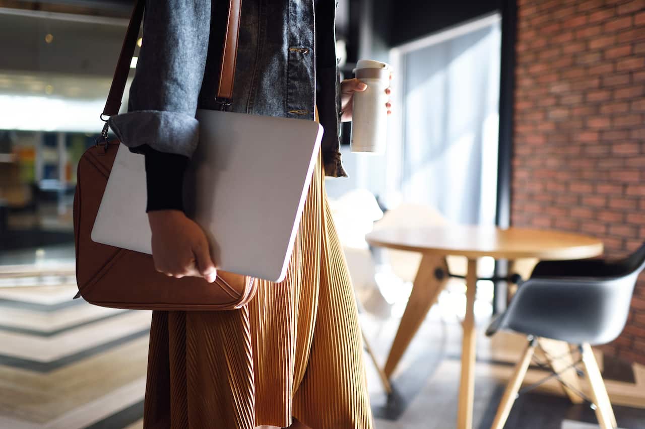 A stock image of a woman carrying laptop, purse and a reusable coffee cup to work.