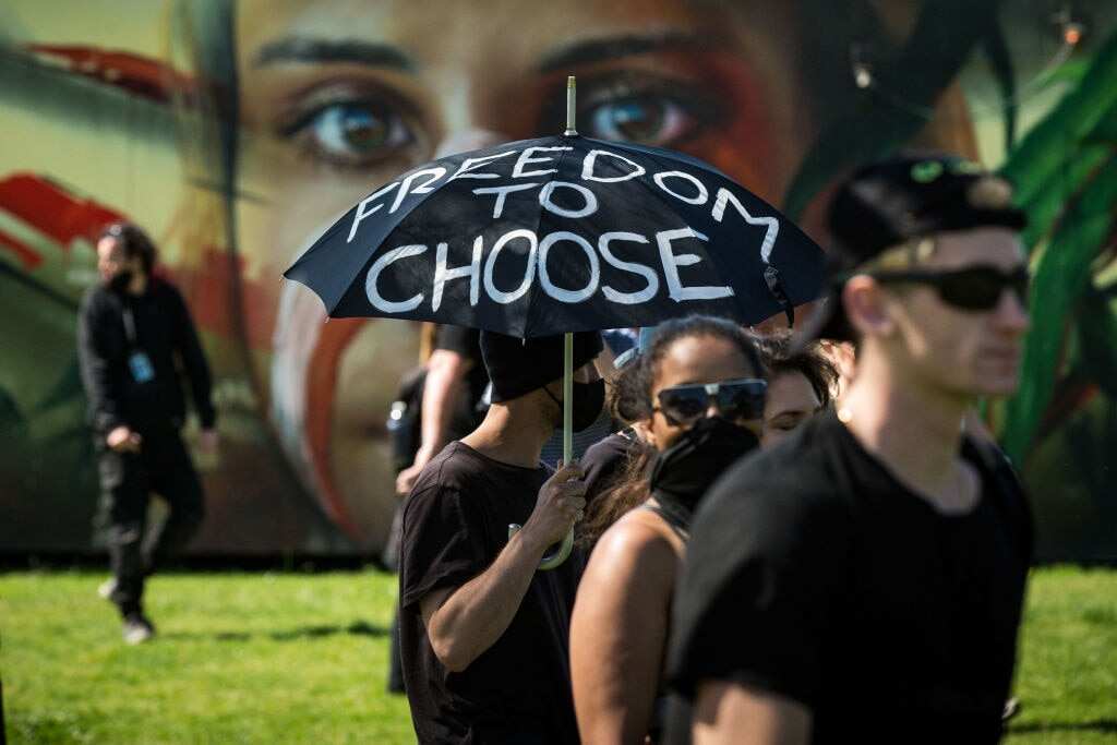 Protesters in Melbourne rally against COVID-19 restrictions. One holds a black umbrella with white writing on it reading 'Freedom to Choose.'
