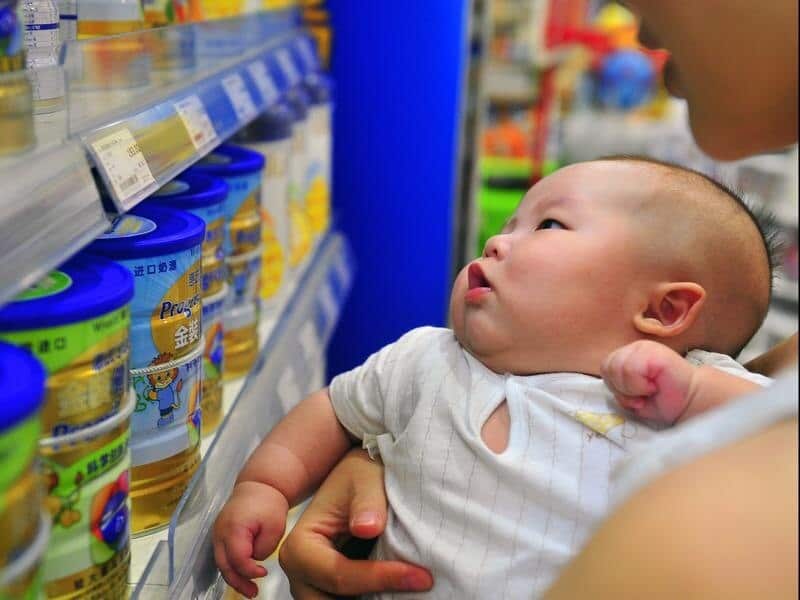 young baby in arms looks at row of infant formula tubs on shop shelf