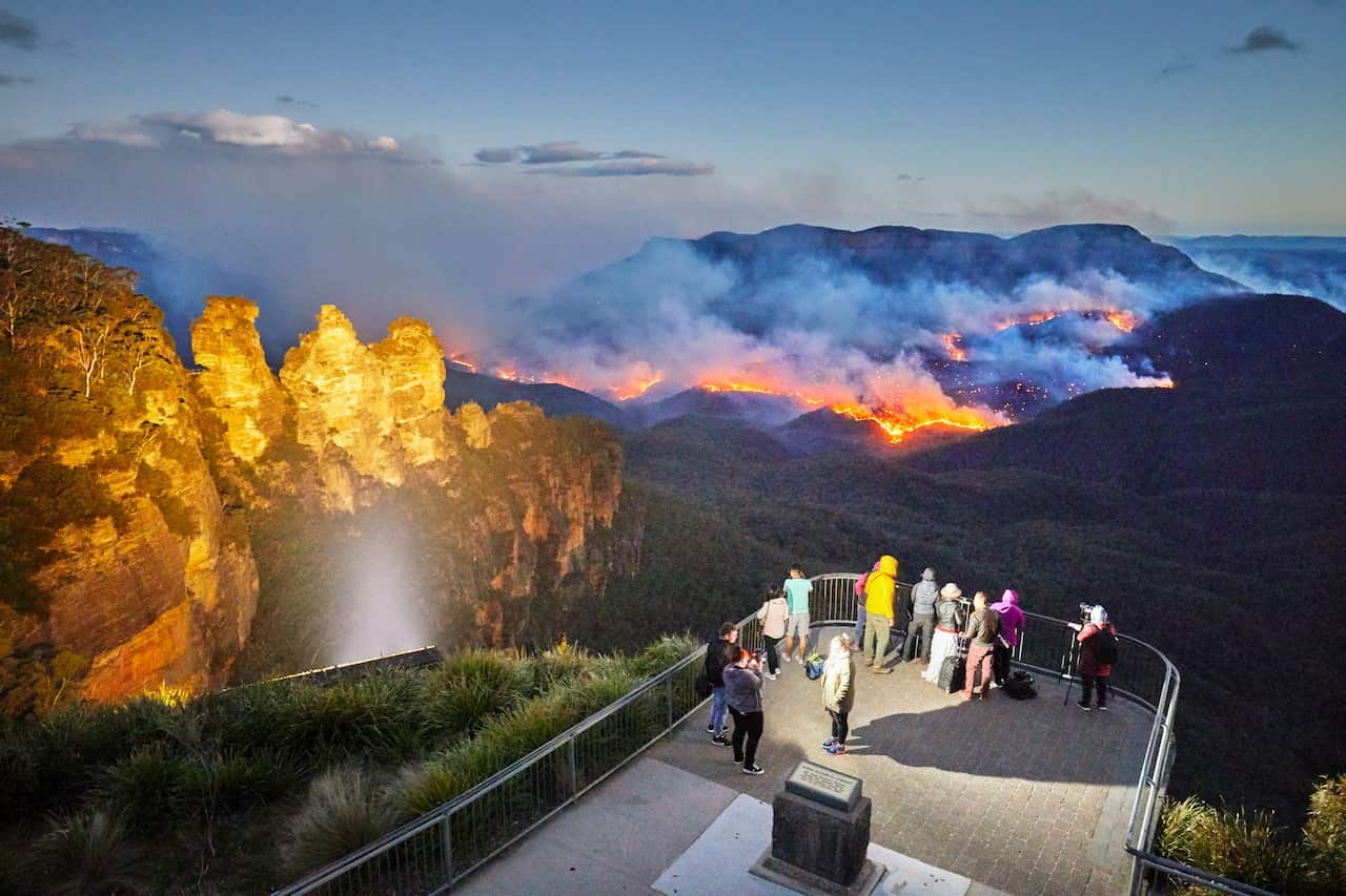 Three Sisters floodlit at dusk, Queen Elizabeth Lookout, viewing platform with tourists watching and photographing fire, bushfire in Jamison Valley, Blue Mountains National Park, Australia