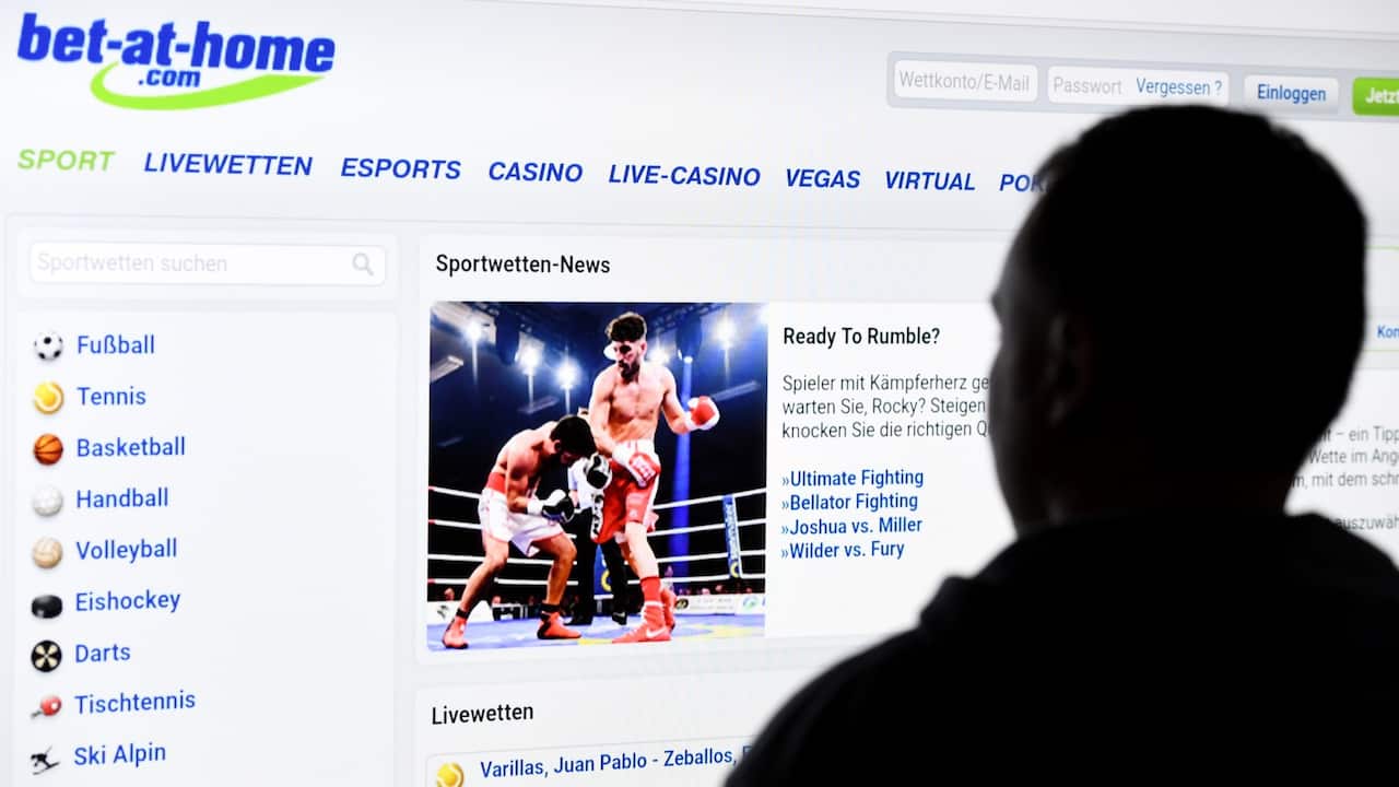 A man stands in front of a screen with a page of a sports betting provider (bet-at-home).