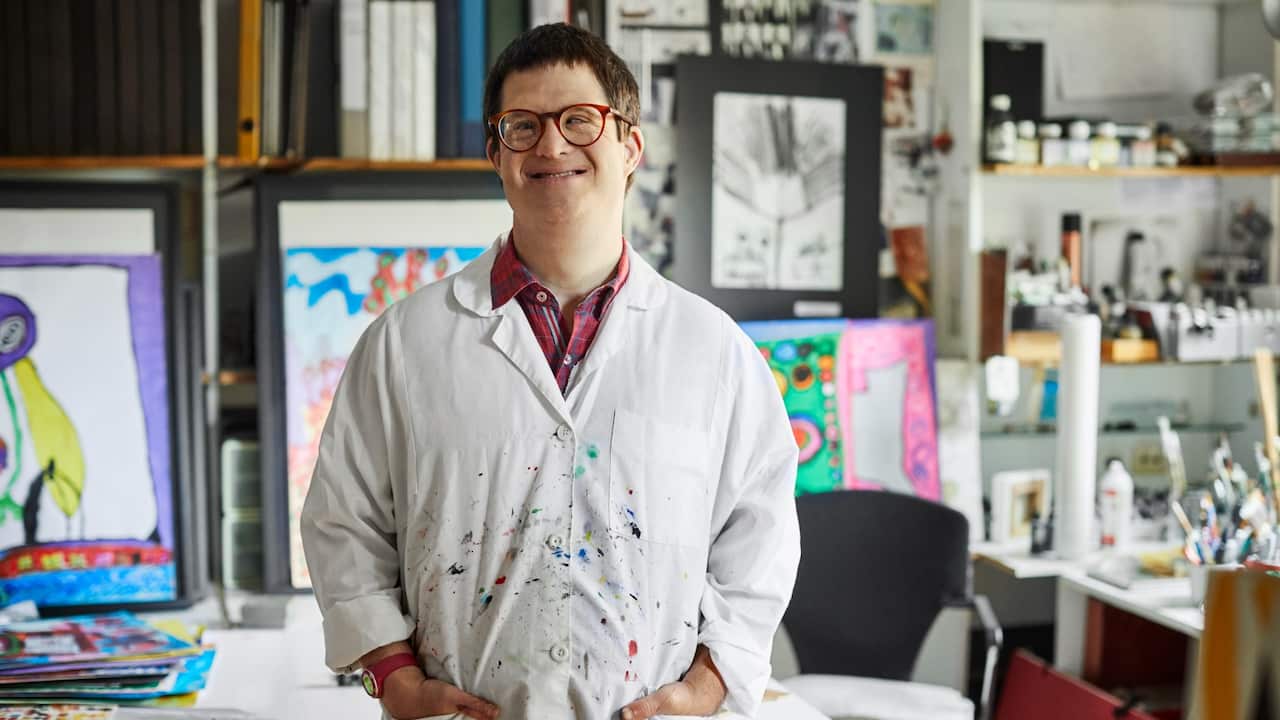 Confident mid adult man with down syndrome in studio. Smiling disabled male artist is wearing apron. He is standing with hands in pockets.