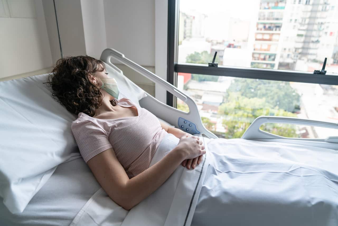 Female hospitalized patient lying down on bed looking through window while wearing a protective face masks