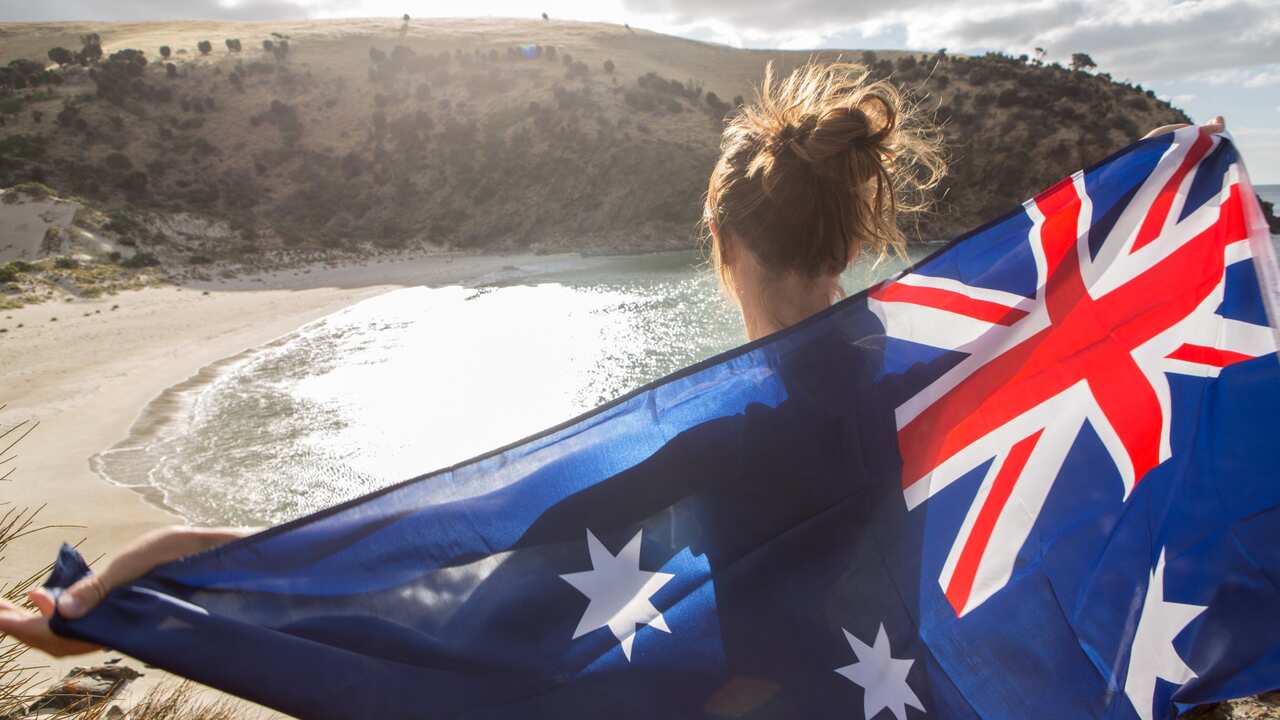 Cheerful young woman stands on a cliff above a beach in Kangaroo Island holding an Australian's flag