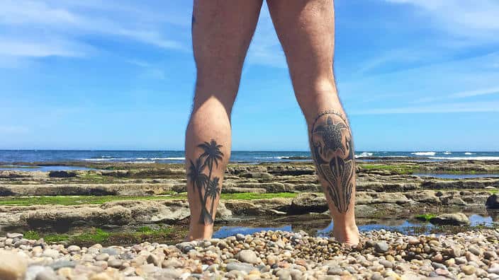 Naked man on his back with tattooed legs looking at the sea on a beach