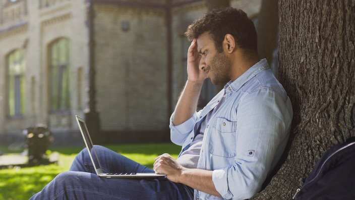 College student sitting under tree with laptop looking upset.