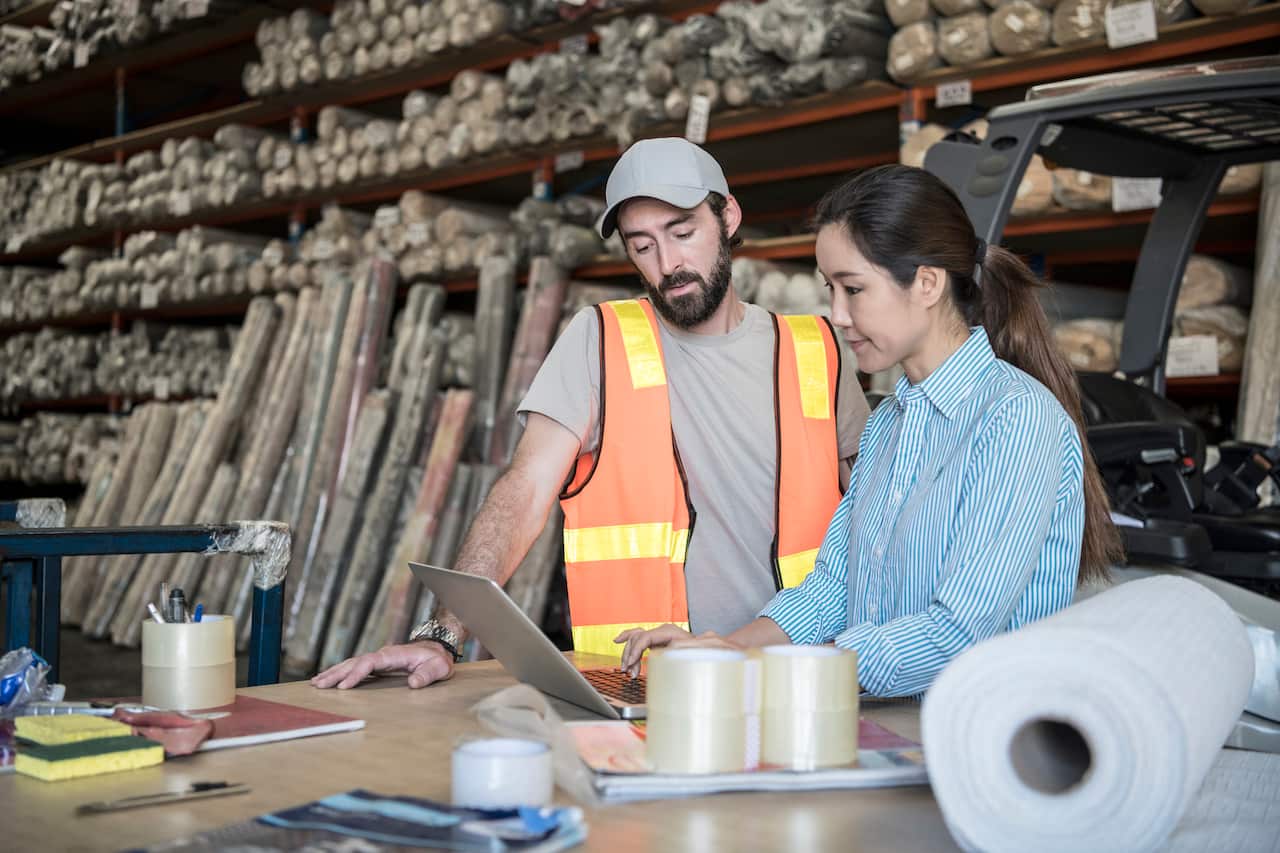 Woman using laptop in warehouse with male employee watching