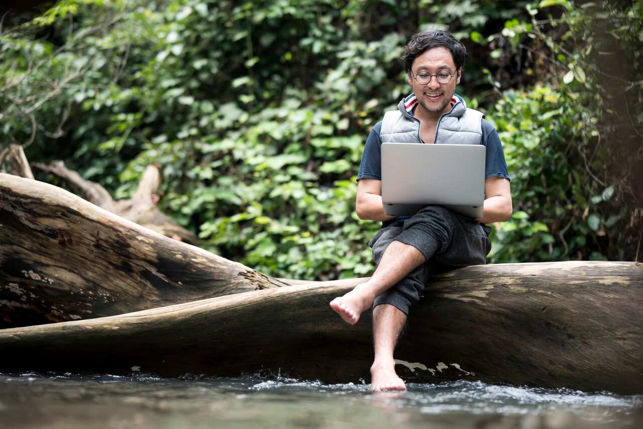 Senior Businessman sitting on a log while reply E-mail or chatting on a laptop on vacation days at a waterfall. Remote Office Anywhere.