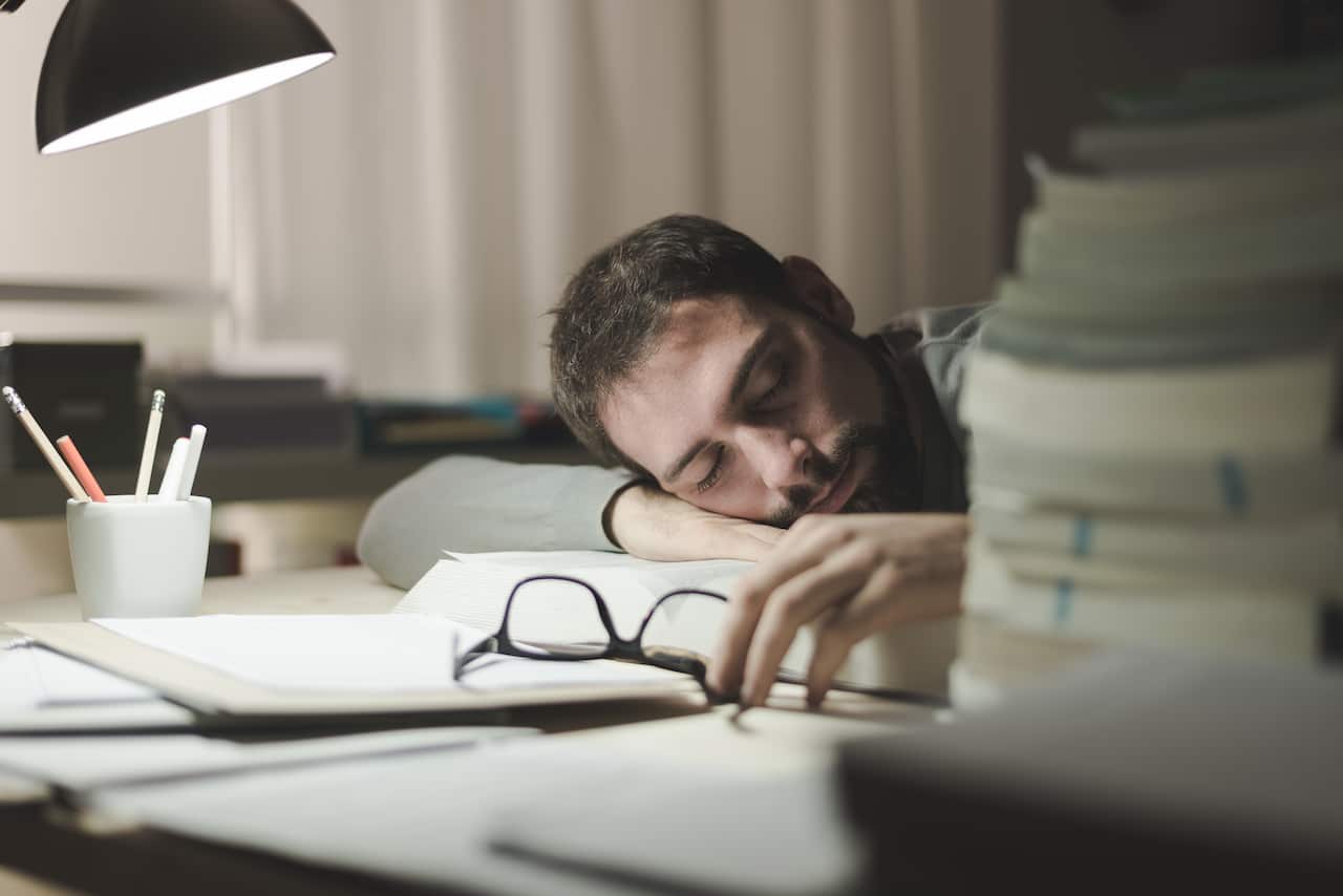 Young man sleeping at his desk late at night, he is leaning on a book and holding glasses, stress and exhaustion concept