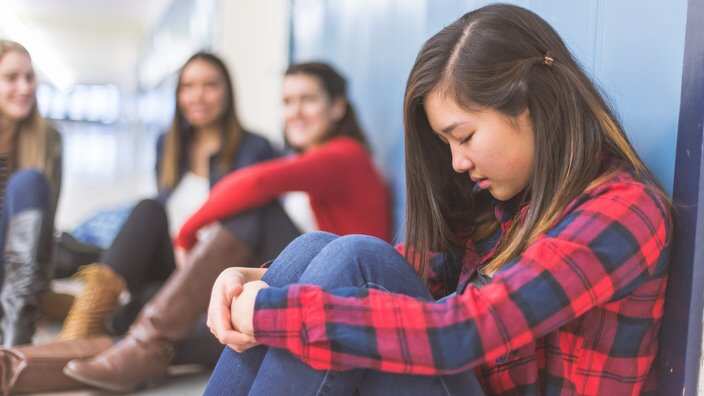 A sad and left-out ethnic high school student leans against a panel of lockers with her eyes closed, head down, and hands clasped to her knees. 
