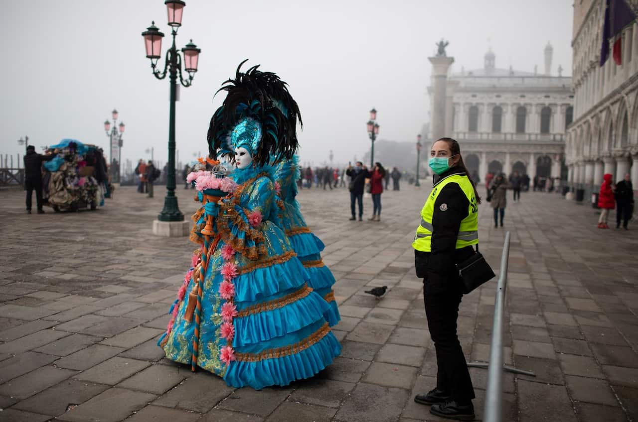 A security guard wearing a protective face mask stand guard as masked and costumed people walk by on the streets of Venice near San Marco square during the Carnival in Venice, Italy, 23 February 2020. 
