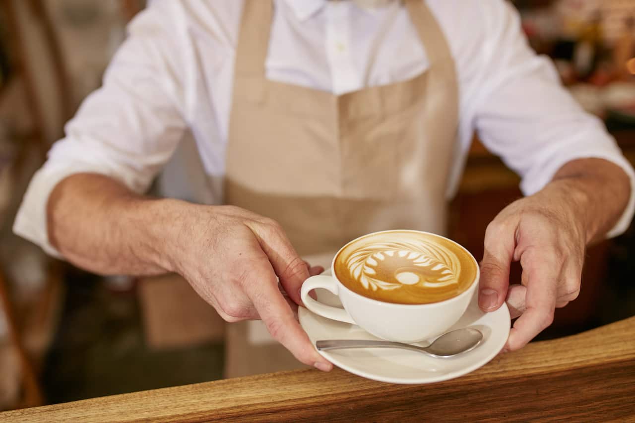 Close-up of man in apron serving coffee while standing in coffee shop. Barista giving you a cup of fresh coffee at cafe.