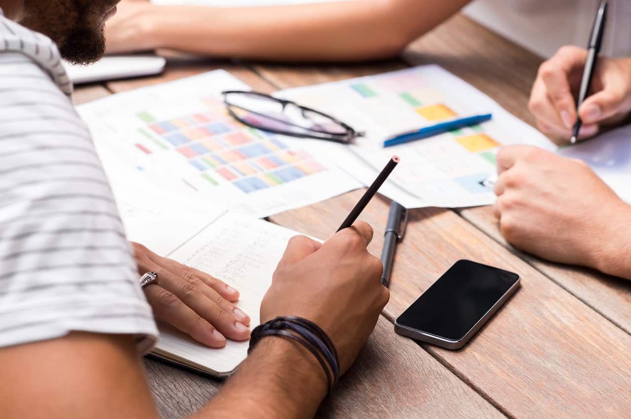 Students discussing the charts and graphs on wooden table. Close up of students hands writing on paper and notebook. Finance students analyzing report of company as assignment.