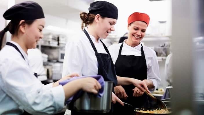 Teacher watching female chef students cooking food in commercial kitchen