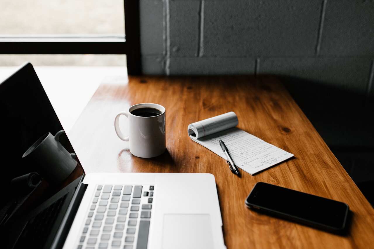 laptop, notebook with pen, mobile phone and cup of coffee seen on desk
