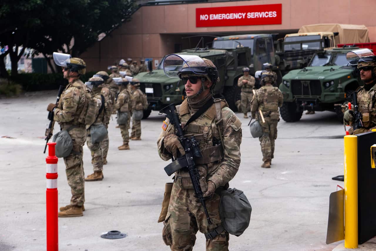 Uniformed national guard troops standing in front of a building, with green vehicles parked behind them.