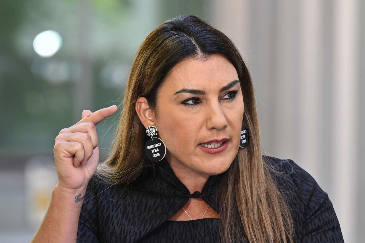 lidia thorpe gestures pointedly, wearing a black top and black earrings bearing the words "Sovereignty never ceded"