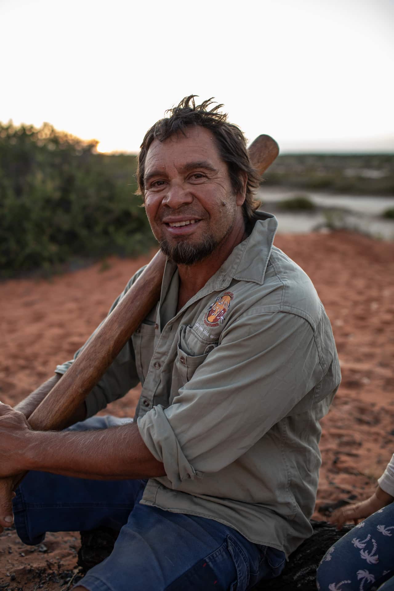 A man holding a didgeridoo, sitting on the ground in the bush.